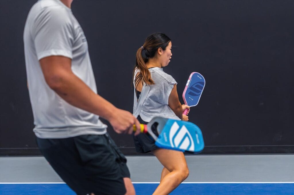 Two people playing pickleball indoors; the woman in the background is about to hit the ball while the man in the foreground holds a paddle.