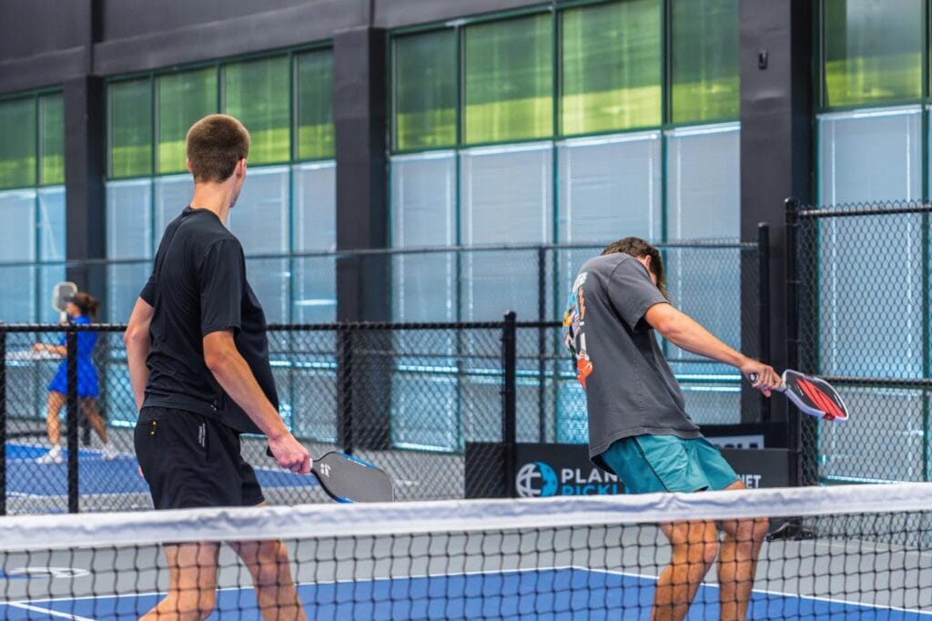 Two men playing pickleball on an indoor court, one hitting the ball with his paddle while the other stands ready.