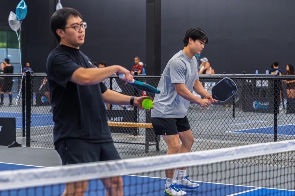 Team Building & Fun: How to Host Your Corporate Pickleball Events 5 Two men on an indoor court play pickleball; one is about to hit the ball with a paddle while the other stands ready behind him.
