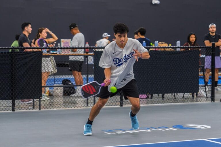 A man wearing a Dodgers jersey plays pickleball indoors, preparing to hit a green ball with his paddle; people watch and socialize in the background.