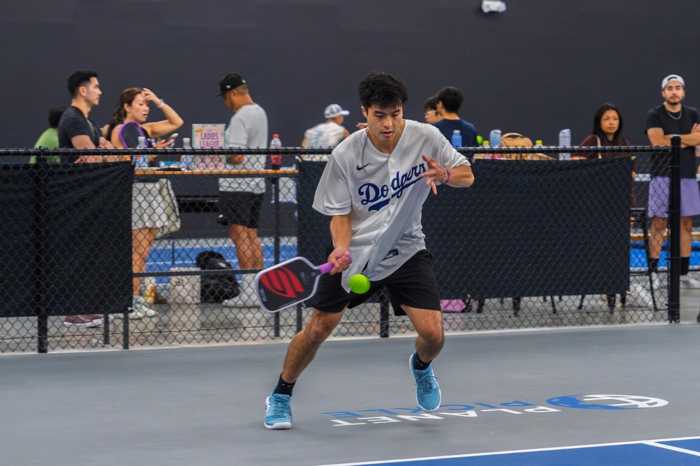 A man wearing a Dodgers jersey plays pickleball indoors, preparing to hit a green ball with his paddle; people watch and socialize in the background.
