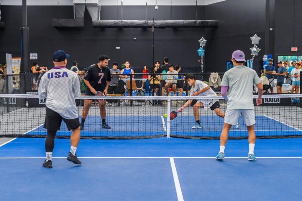 Four men play a doubles pickleball match indoors while spectators watch from behind the court. Balloons and decorations are visible in the background.