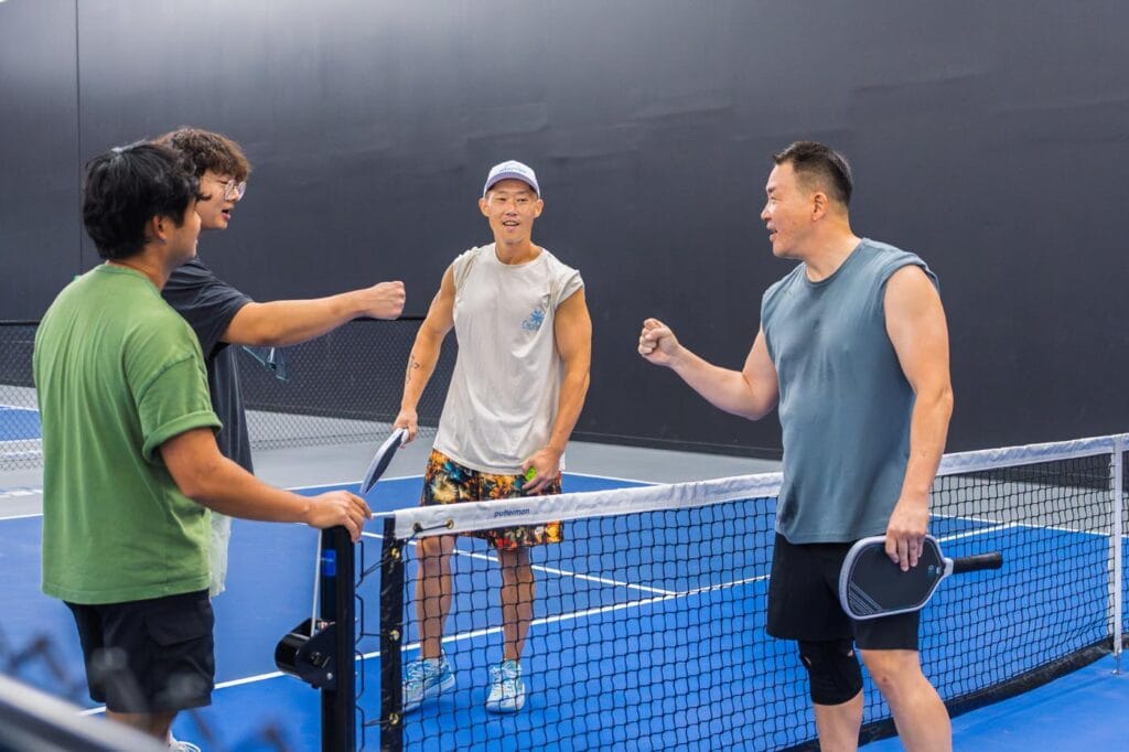Team Building & Fun: How to Host Your Corporate Pickleball Events 3 Four men stand on a blue indoor pickleball court by the net, smiling and giving fist bumps while holding paddles.