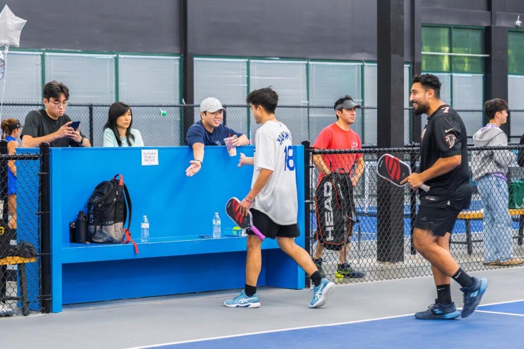 Pickleball for Kids: A Beginner-Friendly Guide for Parents 5 Two men holding paddles walk off a pickleball court while several people stand and sit behind a blue fence, some talking and others using their phones.