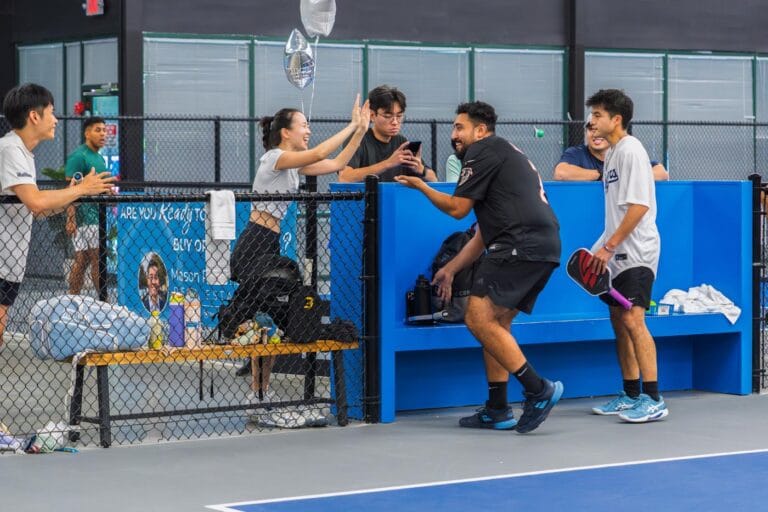 A group of people socialize and laugh near a pickleball court, with some sitting on a bench and others standing, holding paddles and water bottles.
