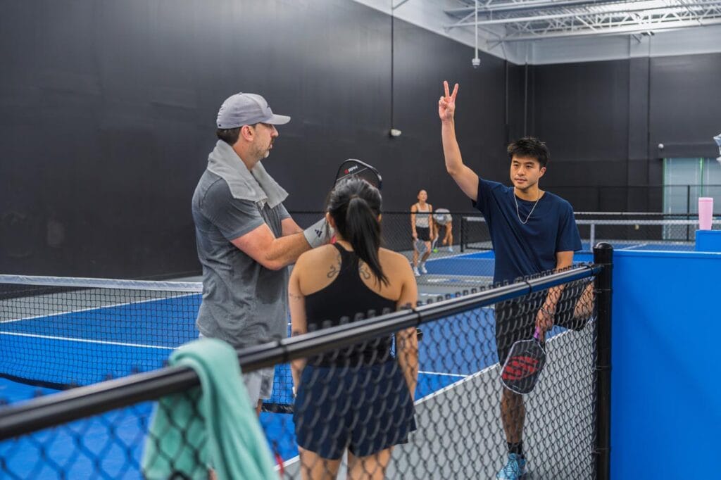 Three people stand near a net on an indoor tennis court; one man holds up two fingers, while the others hold rackets and talk.