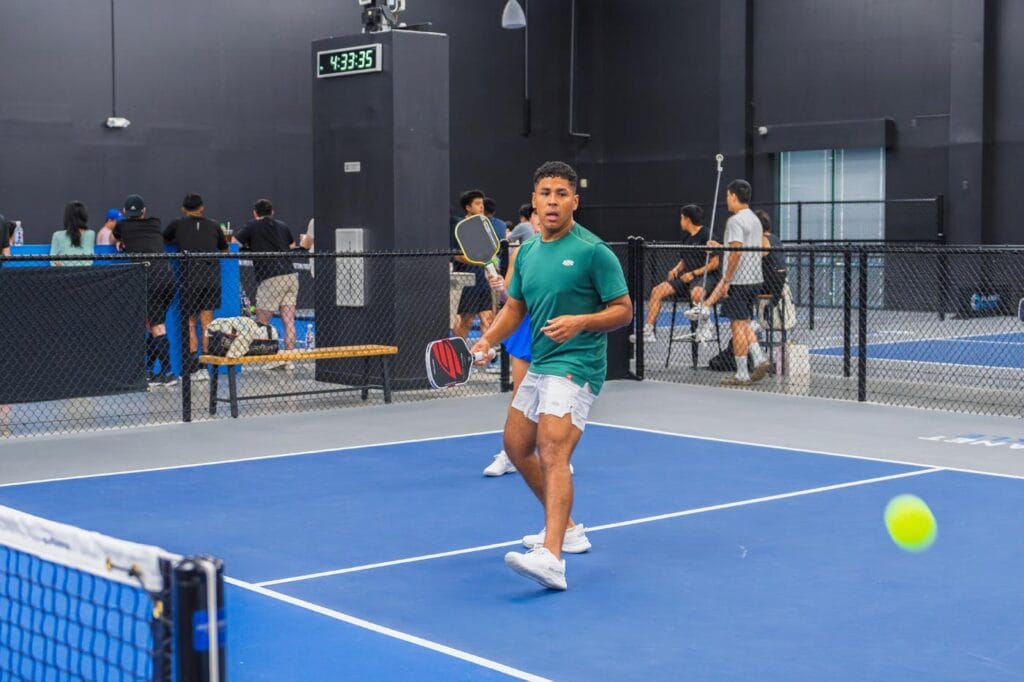 Team Building & Fun: How to Host Your Corporate Pickleball Events 1 Two people play pickleball on an indoor court, with one person preparing to hit the ball. Several others watch or wait behind a fence in the background.