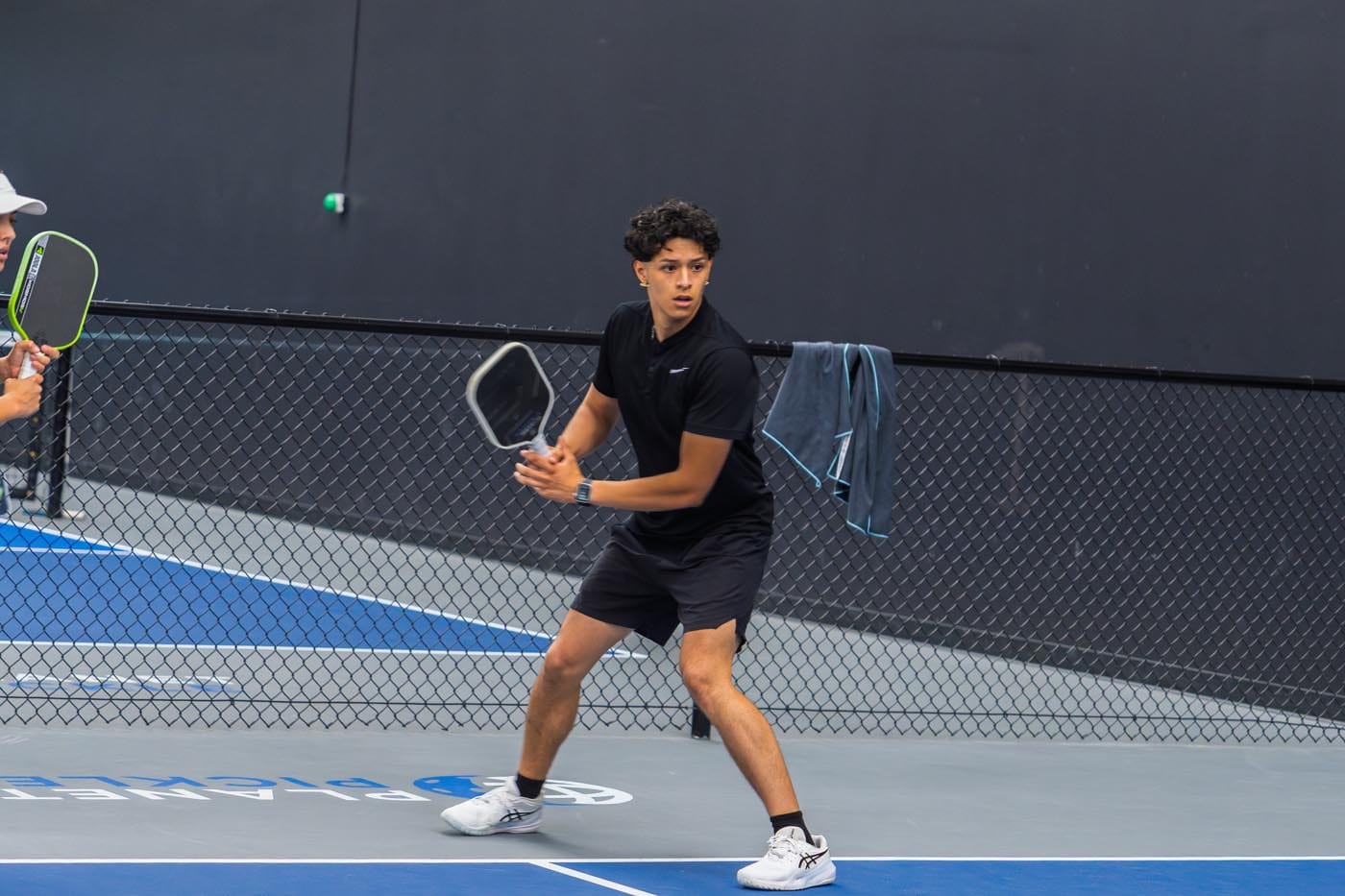 A young man in black sportswear prepares to hit a ball with a paddle on an indoor pickleball court.