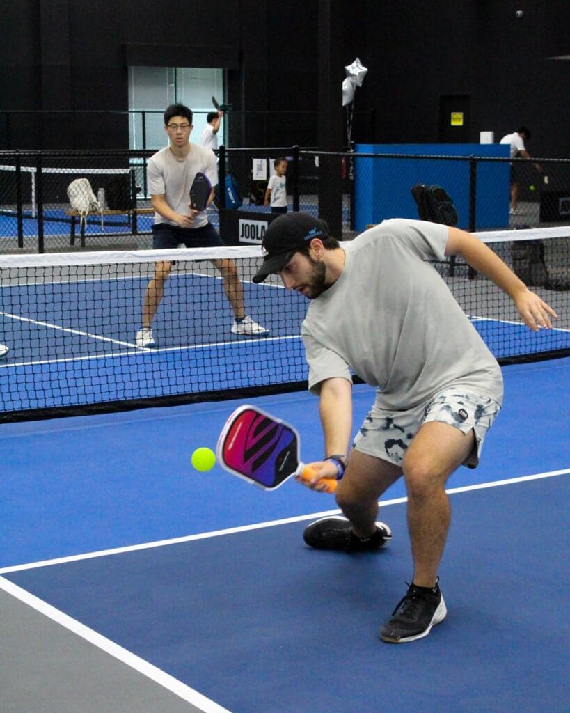 Two men play pickleball on an indoor court; one lunges forward to hit the ball with his paddle while the other stands ready near the baseline.