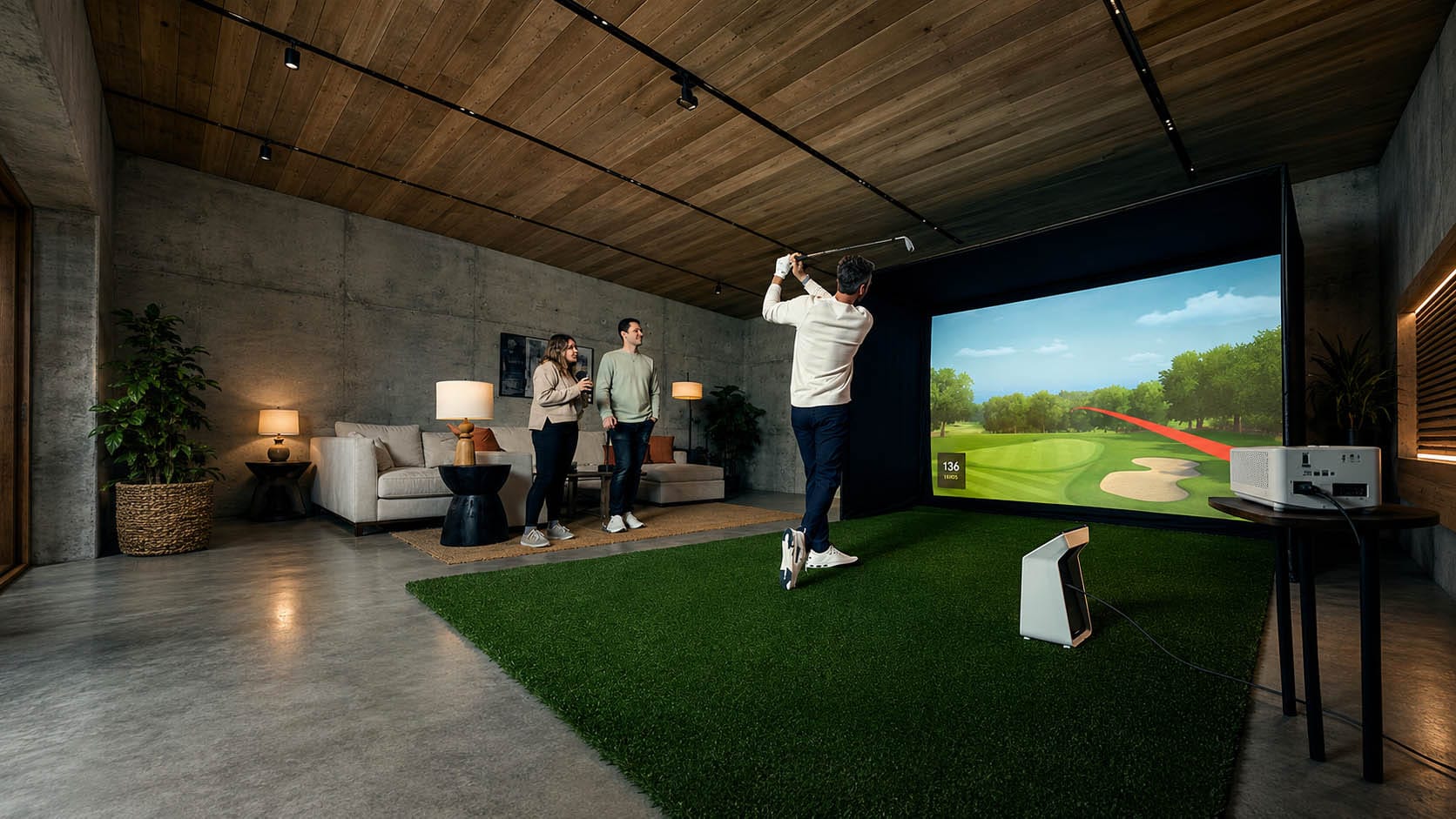 Person swings a golf club on an indoor golf simulator while two people watch from a couch in a modern living room with concrete walls and wood ceiling.