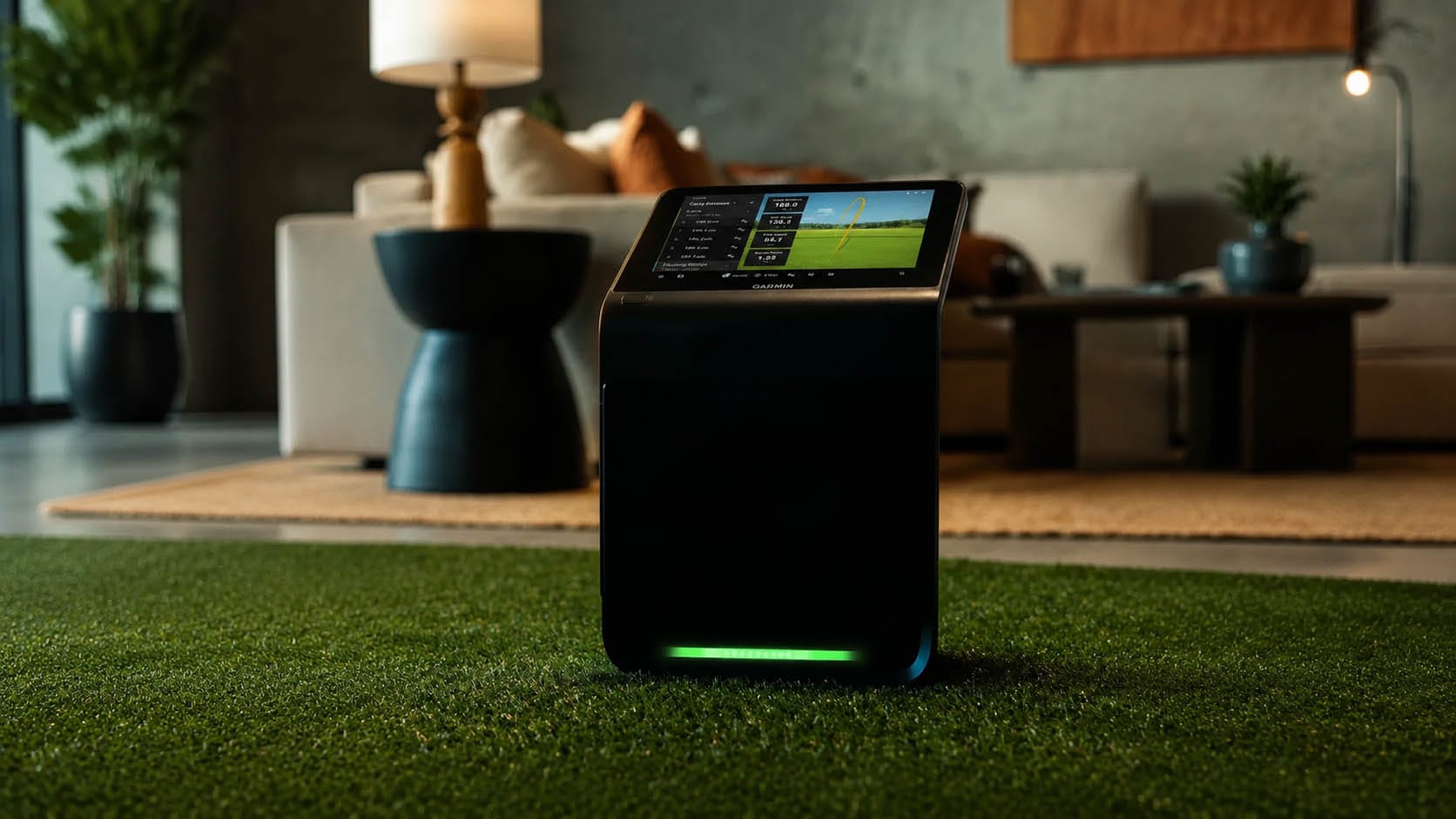 A golf simulator device with a screen displays stats, positioned on artificial turf in a modern living room with a sofa and a coffee table in the background.