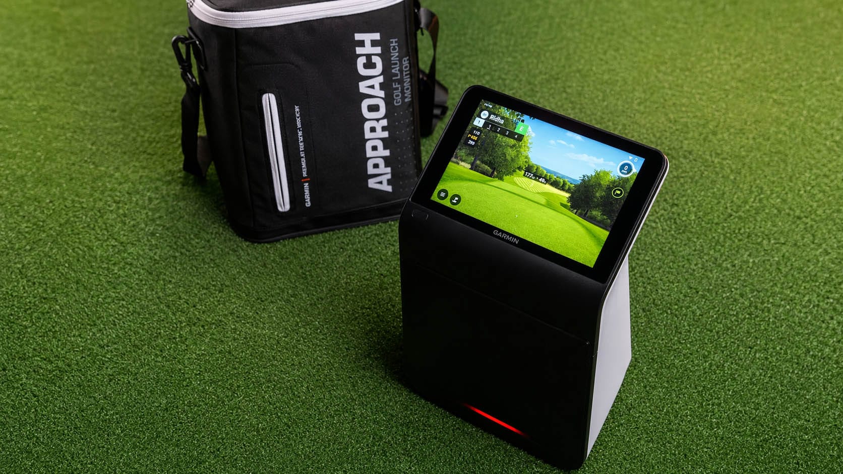 A Garmin golf launch monitor with a touchscreen display sits on artificial turf next to a black carrying bag labeled “Approach.”.