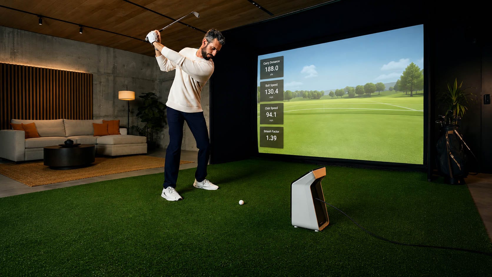 A man swings a golf club in an indoor golf simulator room with data projected on a large screen.
